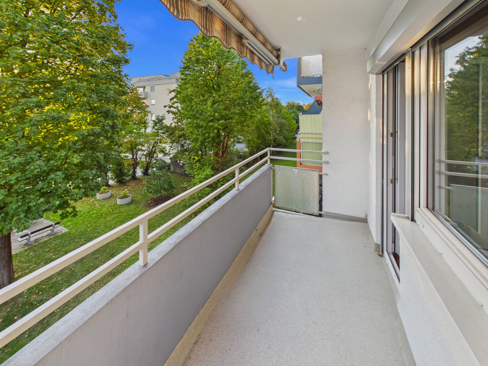 Balkon mit Markise, weißem Geländer und Blick auf grüne Bäume im Garten.