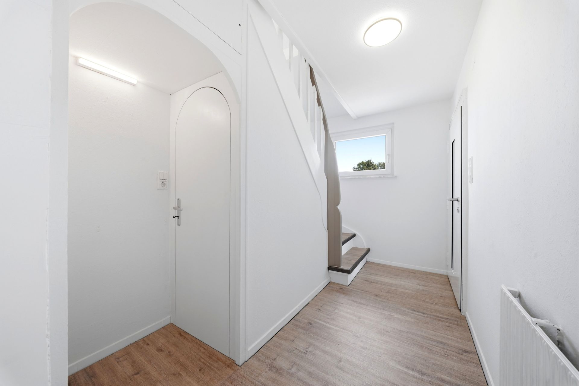 Heller Flur mit weißer Tür, Holztreppe und Fenster, Boden in Holzoptik.