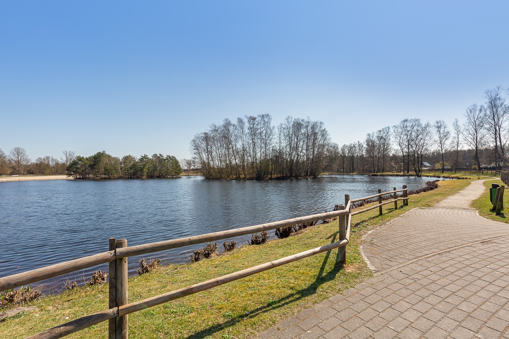 See mit Uferweg, Holzgeländer und Bäumen im Hintergrund bei klarem Himmel.