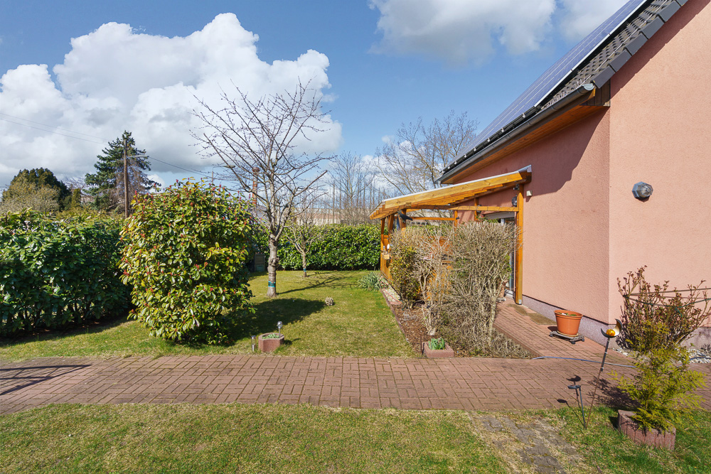 Garten mit Rasen, Sträuchern, Bäumen und Terrasse am Haus mit Solarpaneelen.