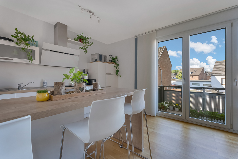Moderne Küche mit Bartresen, weißen Stühlen, Pflanzen und großer Fensterfront mit Blick nach draußen.