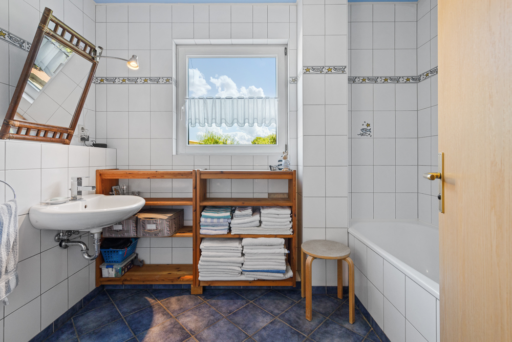 Helles Badezimmer mit Waschbecken, Badewanne, Regal mit Handtüchern und Fenster.