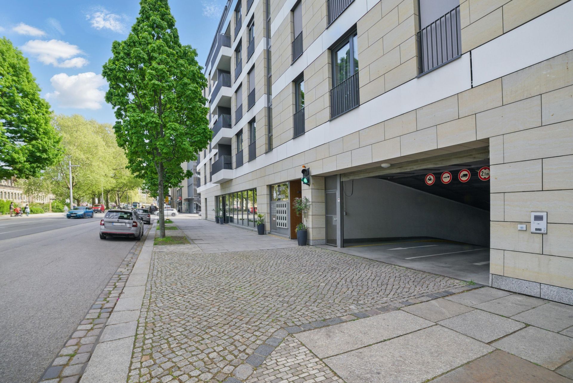 Moderne Wohnhausfassade mit geöffneter Tiefgarageneinfahrt und Bäumen am Straßenrand.