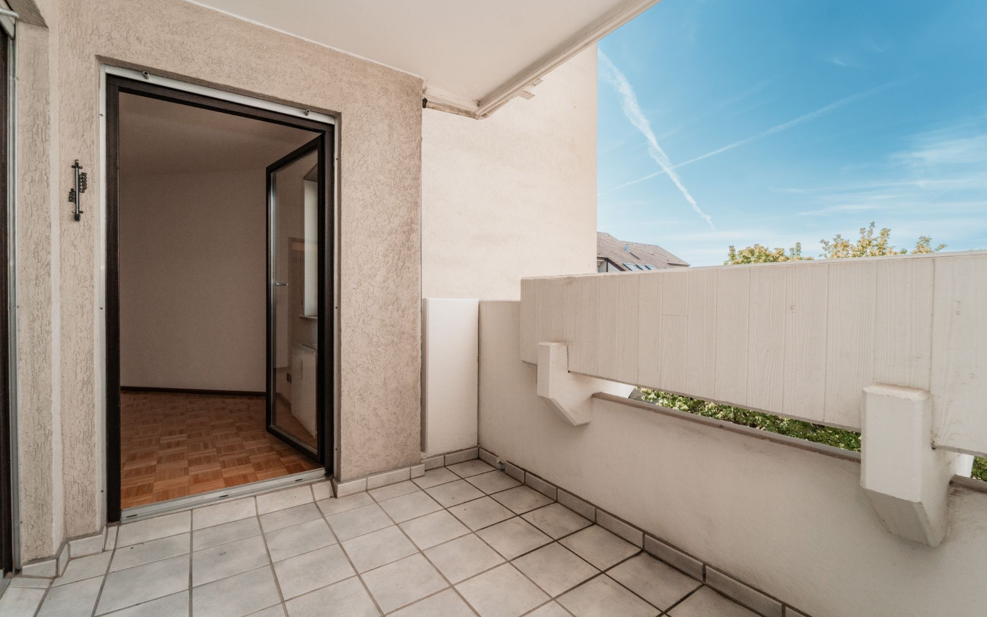 Überdachter Balkon mit Fliesenboden und offener Tür zu einem Innenraum, blauer Himmel sichtbar.