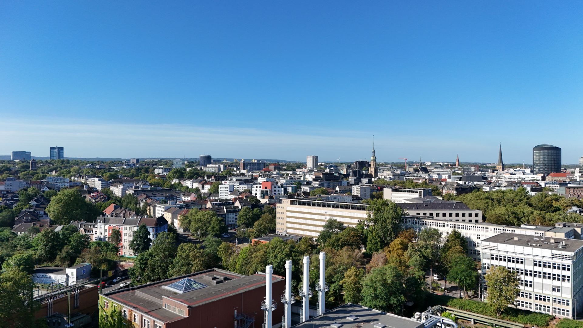 Stadtpanorama mit vielen Gebäuden, Bäumen und blauem Himmel im Hintergrund.