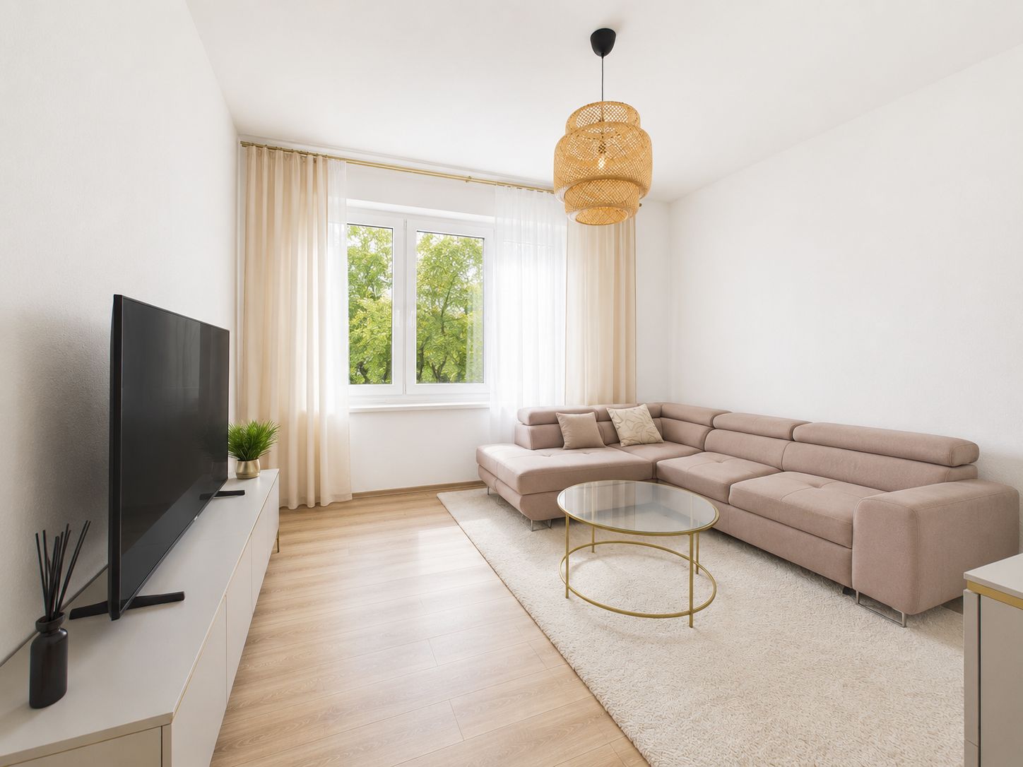 Helles Wohnzimmer mit beiger Eckcouch, Glastisch und großem Fernseher vor Fenster.