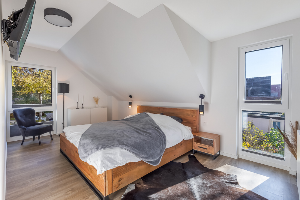 Modernes Schlafzimmer mit großem Fenster, Holzbett, Sessel und Kuhfell-Teppich auf Holzboden.