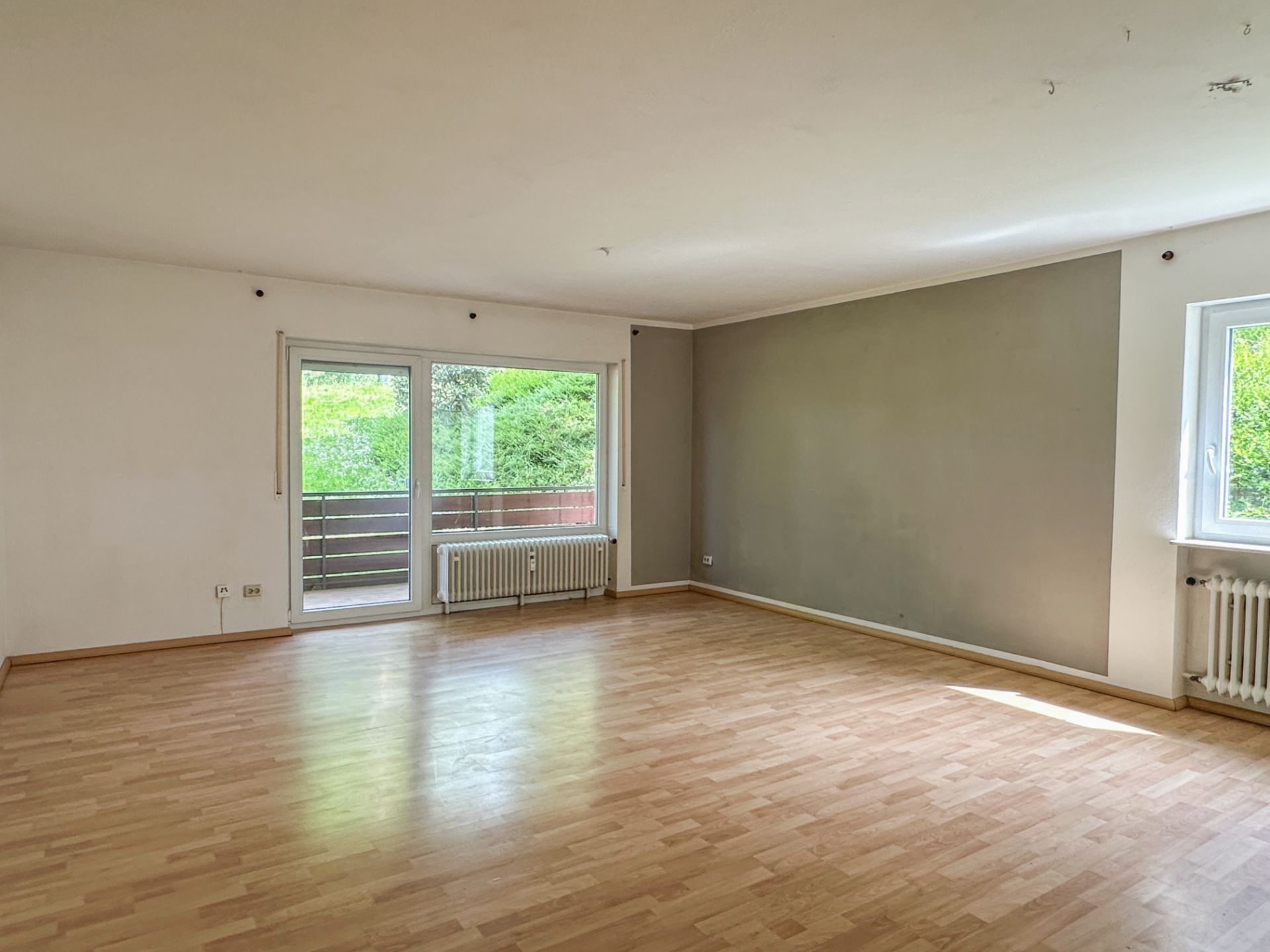 Helles, leeres Zimmer mit Holzboden, zwei Heizkörpern und großen Fenstern.