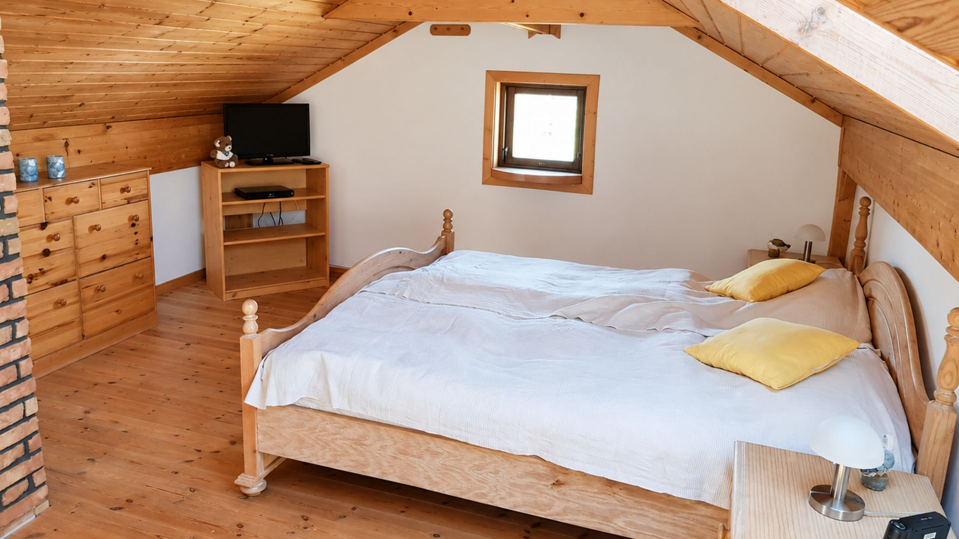 Schlafzimmer mit Doppelbett, Holzdecke, Nachttisch, Kommode und kleinem Fenster.