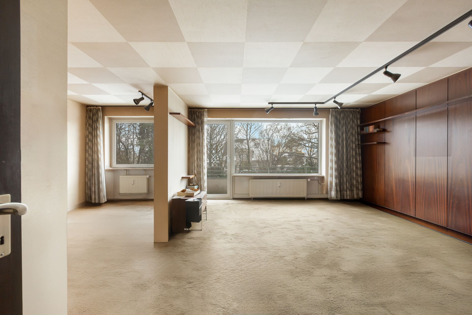 Großes, helles Wohnzimmer mit Teppichboden, Holzdecke und großen Fenstern mit Vorhängen.