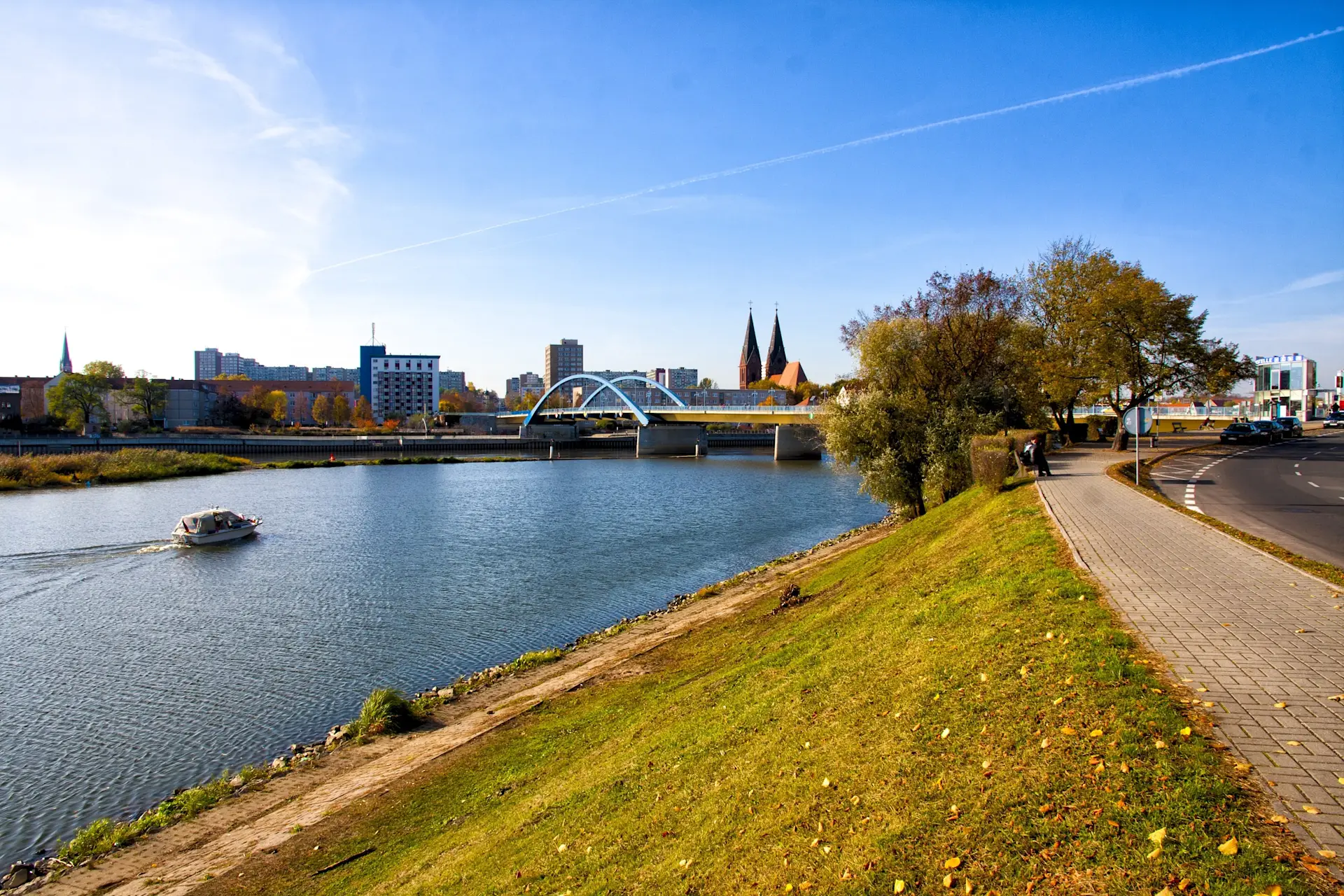 Fluss mit Brücke, Boot und Uferweg, im Hintergrund Stadt und Bäume.