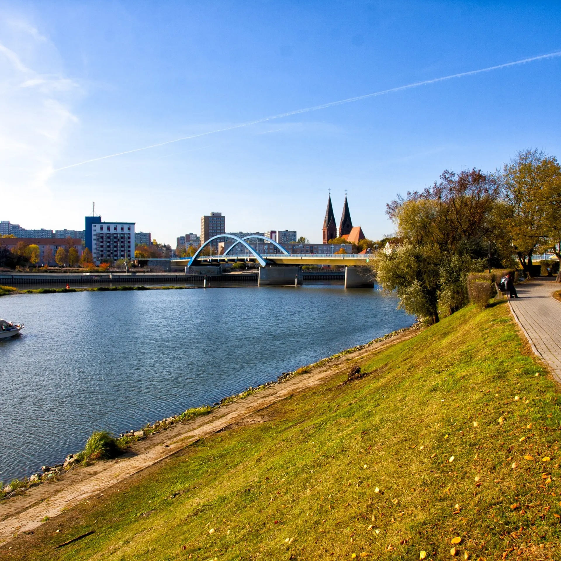 Fluss mit Brücke, Boot und Uferweg, im Hintergrund Stadt und Bäume.