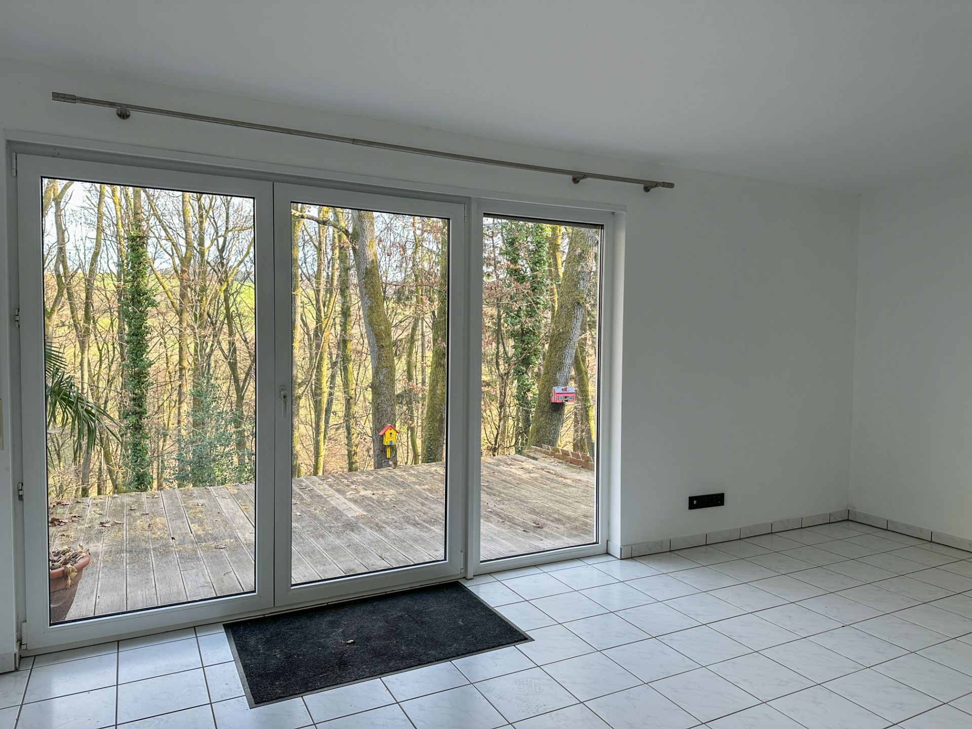 Heller Raum mit weißen Fliesen, großer Glasfront und Blick auf eine Holzterrasse und Bäume.