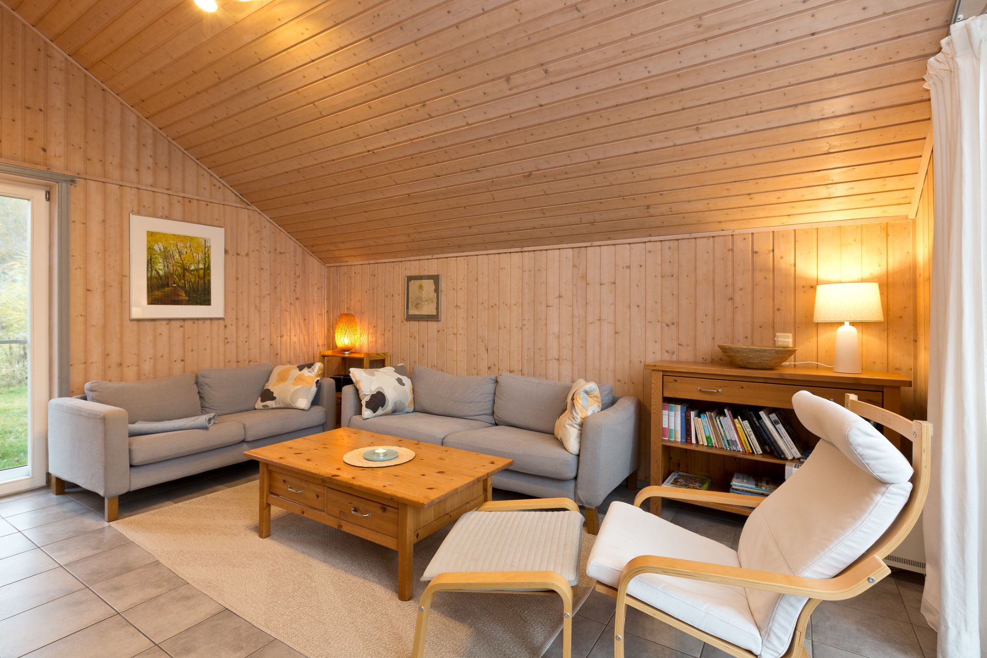 Gemütliches Wohnzimmer mit Holzwänden, zwei Sofas, Sessel, Couchtisch und Bücherregal mit Lampe.