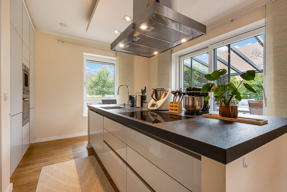 Moderne Küche mit Kochinsel, Pflanzen und Küchengeräten, Blick nach draußen durch große Fenster.