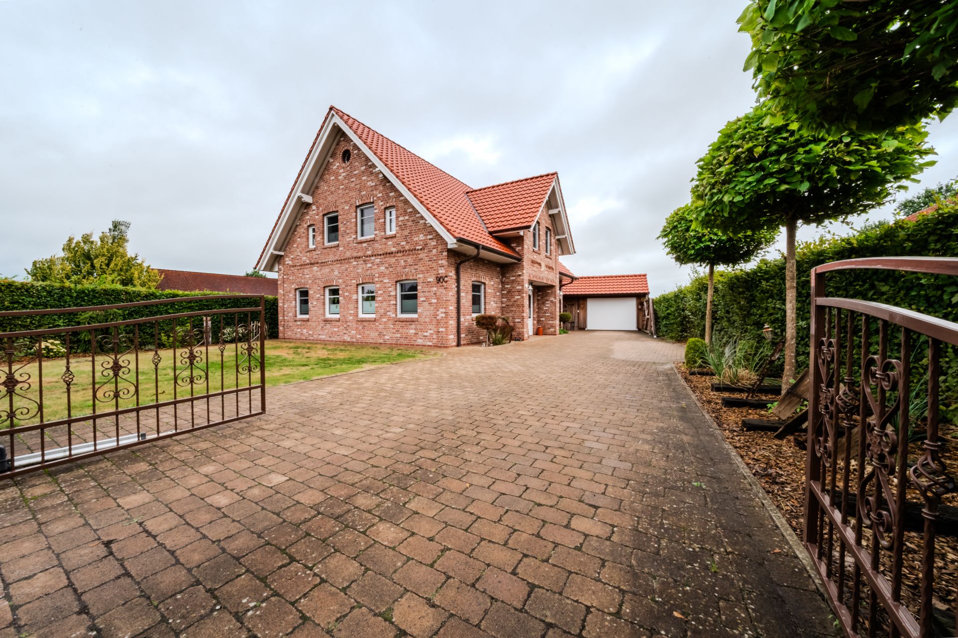 Backsteinhaus mit rotem Dach, gepflasterter Einfahrt und grünem Garten, gesehen durch ein offenes Tor.