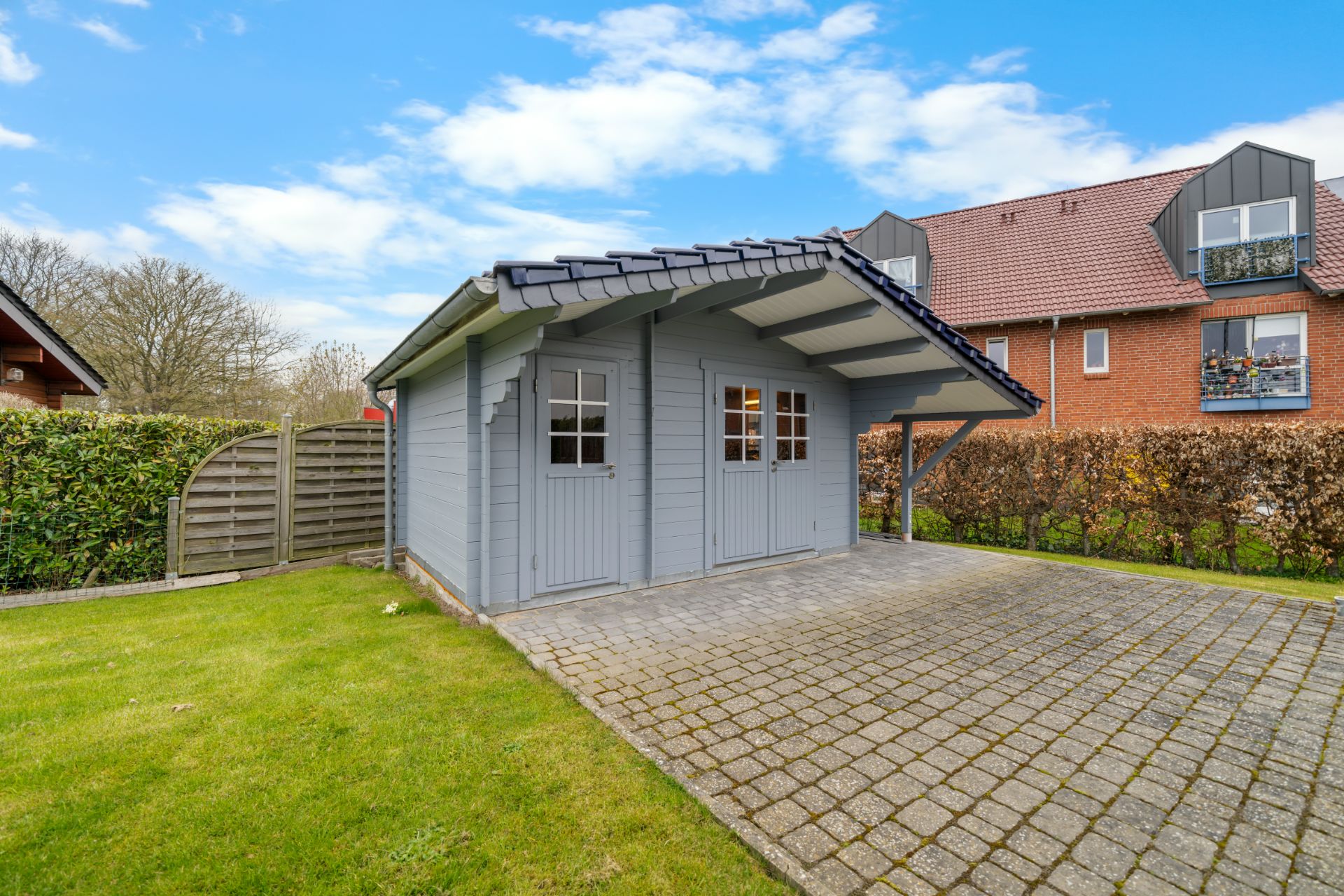 Gartenhaus mit überdachtem Bereich auf gepflastertem Platz und Rasenfläche im Garten.