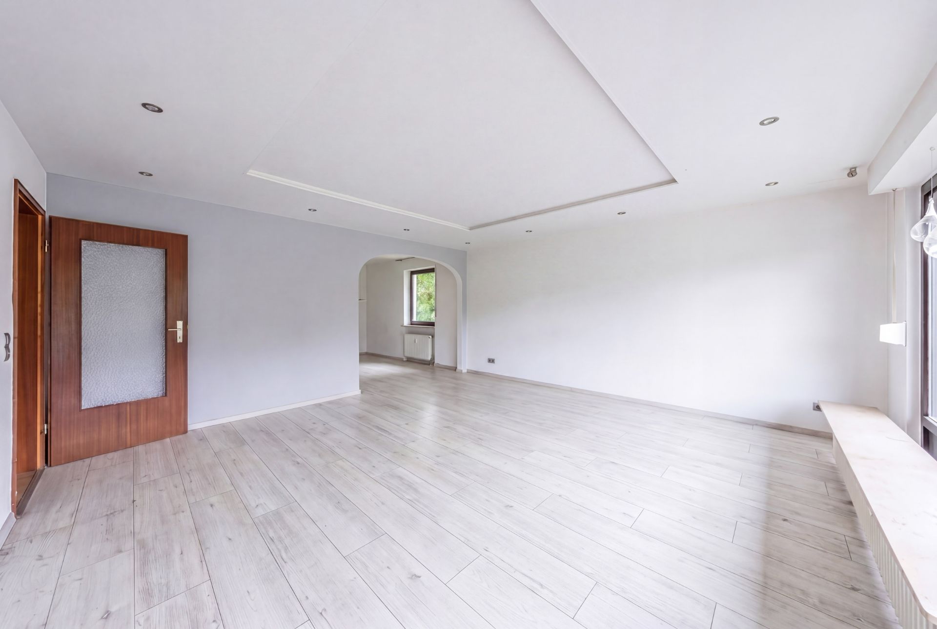 Helles, leeres Wohnzimmer mit großen Fenstern, Holztür und hellem Boden.