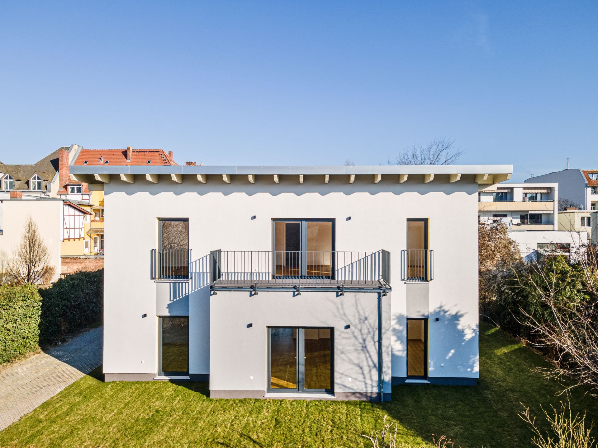 Modernes weißes Wohnhaus mit Balkon und großen Fenstern, umgeben von grünem Garten.