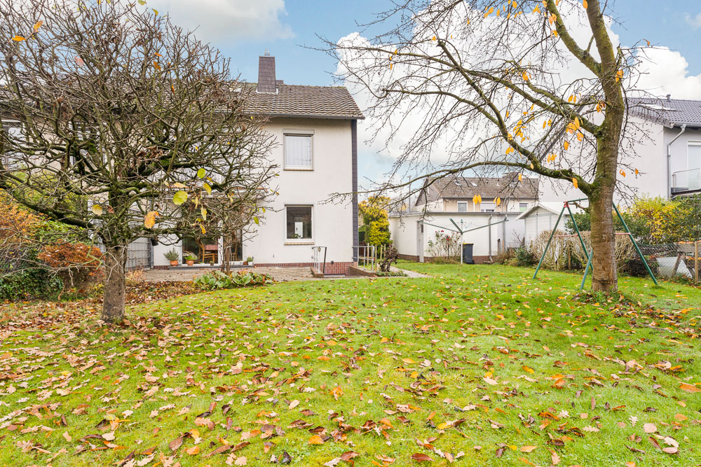 Garten mit Rasen, zwei Bäumen und Blick auf ein helles Wohnhaus im Hintergrund.