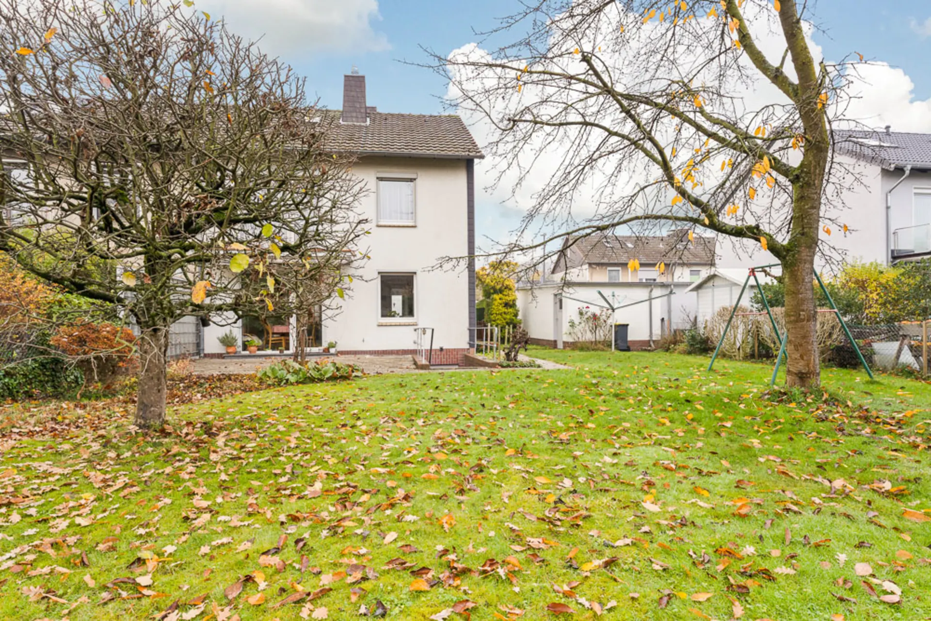 Garten mit Rasen, zwei Bäumen und Blick auf ein helles Wohnhaus im Hintergrund.