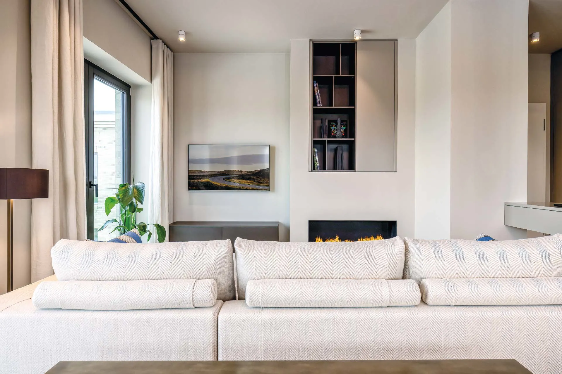 Helles Wohnzimmer mit cremefarbenem Sofa, Kamin, Regal, großem Fenster und grüner Pflanze.