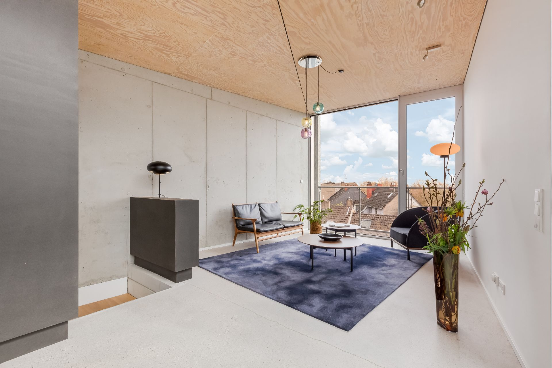 Modernes Wohnzimmer mit großen Fenstern, blauem Teppich, Sitzmöbeln und Zimmerpflanzen.
