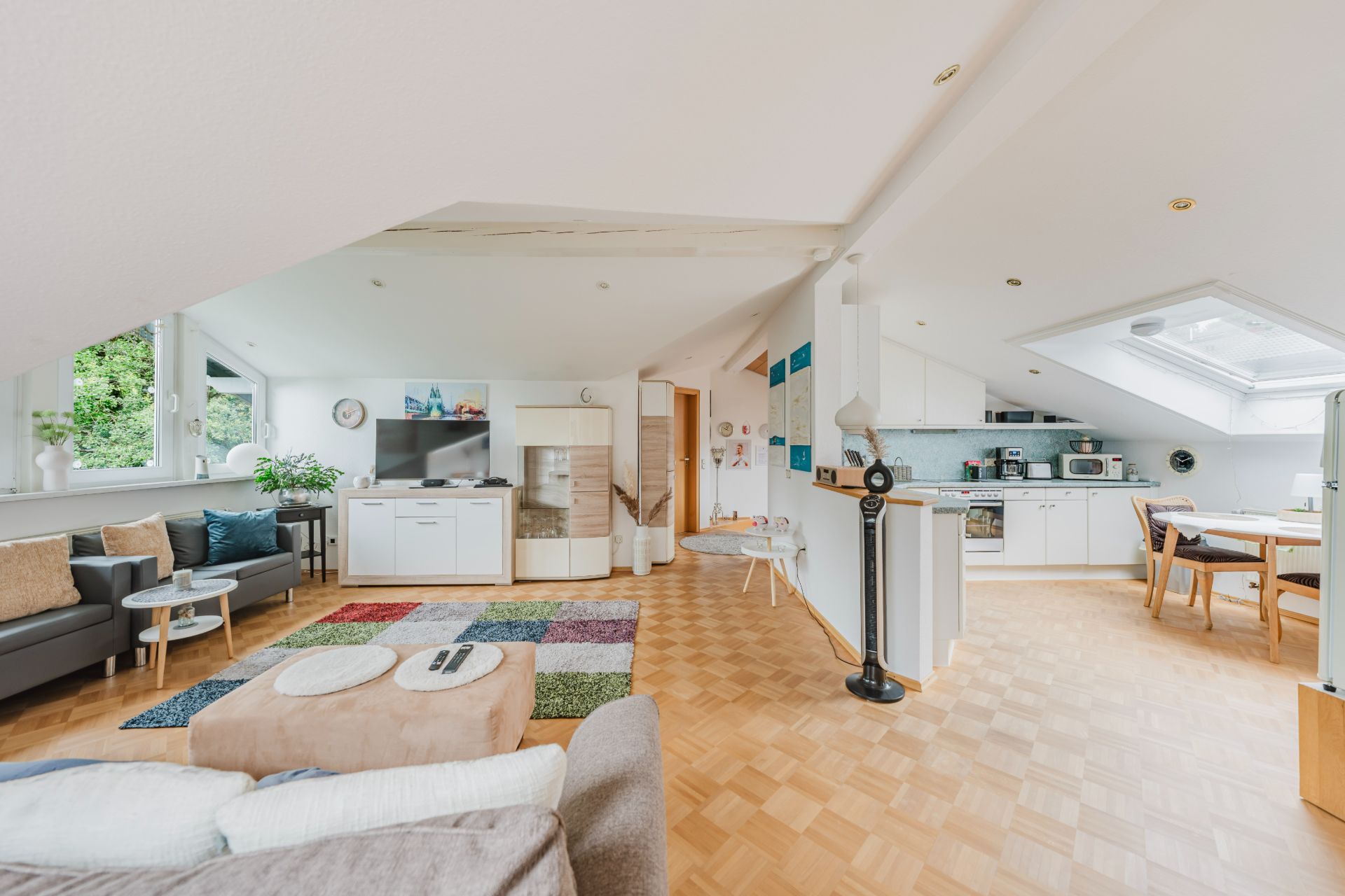Helles Wohnzimmer mit Sofa, Fernseher, bunter Teppich, angrenzender Küche und Essbereich.