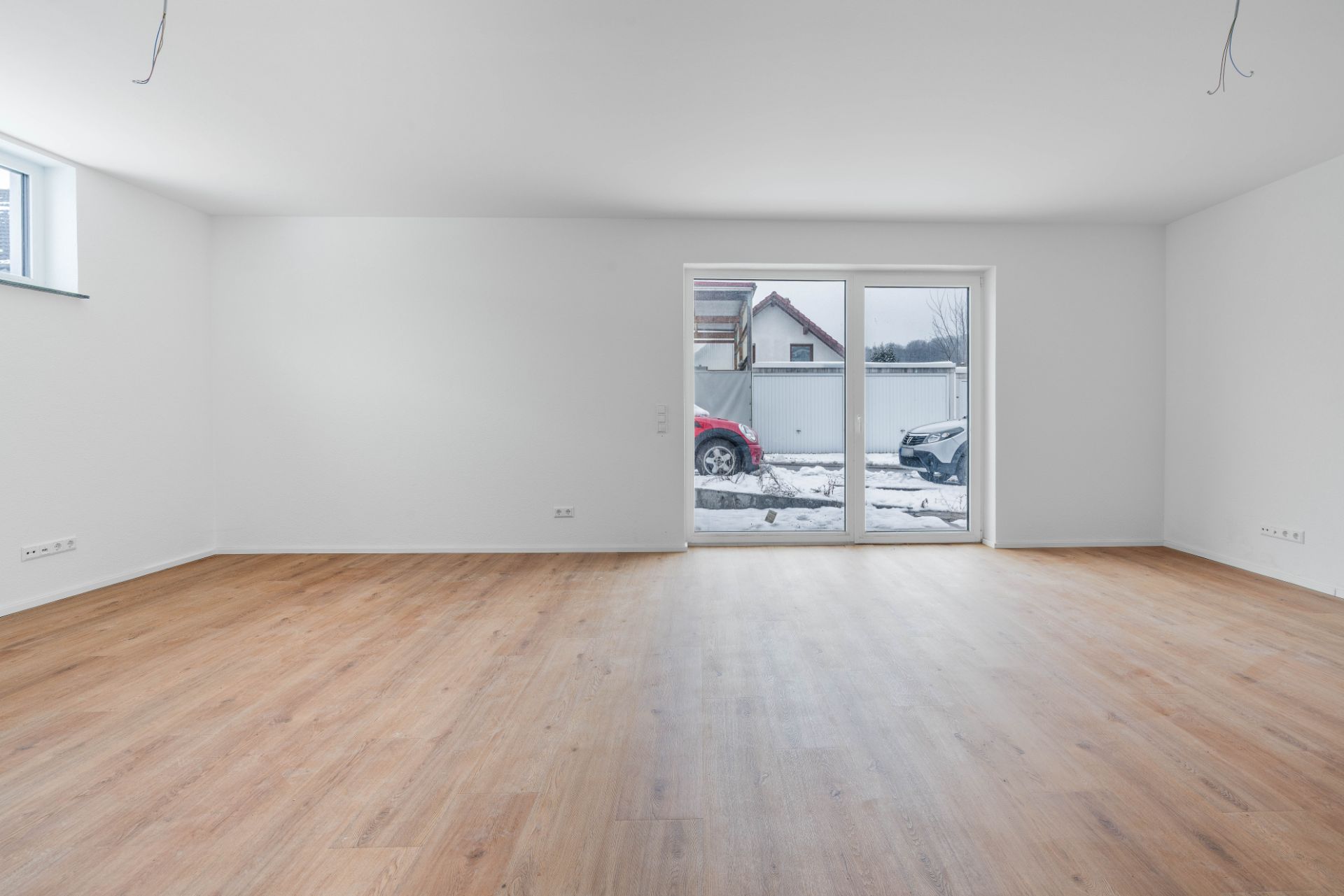 Leeres Zimmer mit Holzfußboden, weiß gestrichenen Wänden und großer Glastür zum Garten.