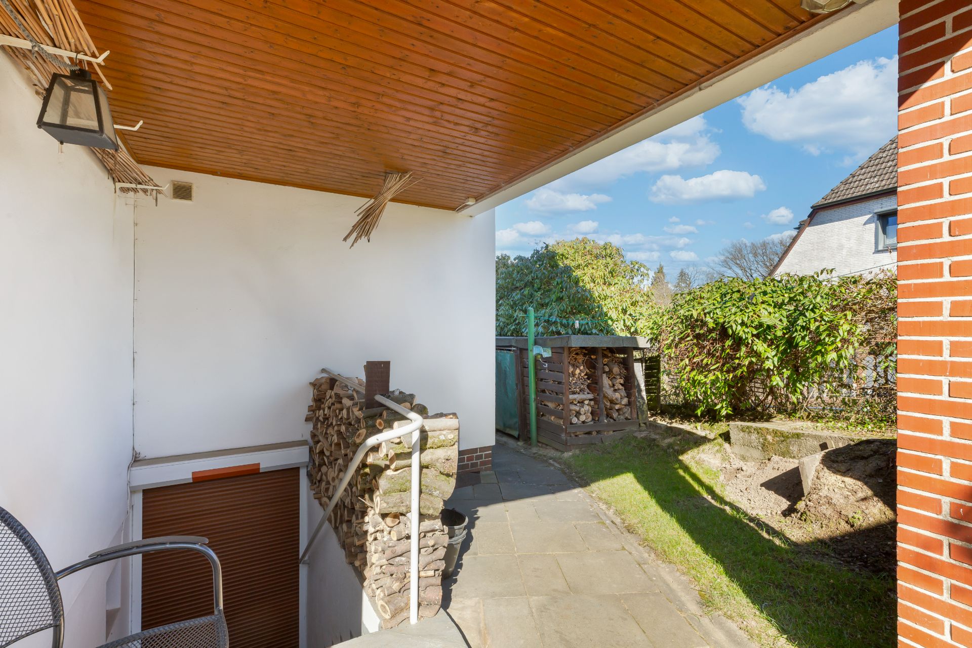Überdachte Terrasse mit Stühlen, Holzstapel und Zugang zum Garten.