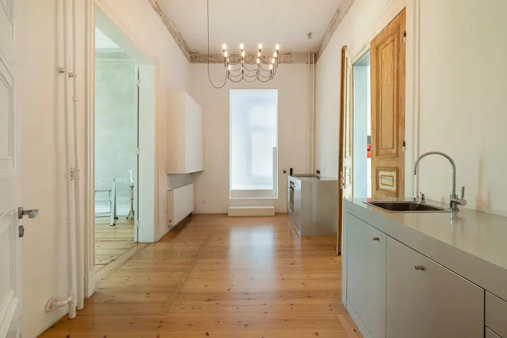 Heller Raum mit Holzboden, moderner Küchenzeile, großem Fenster und Kronleuchter.