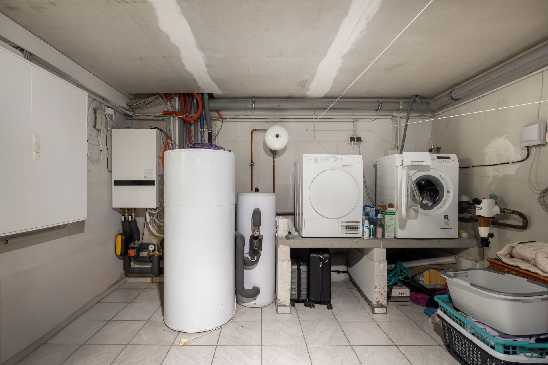 Waschküche mit Waschmaschine, Trockner, Boiler und Regalen auf hell gefliestem Boden.