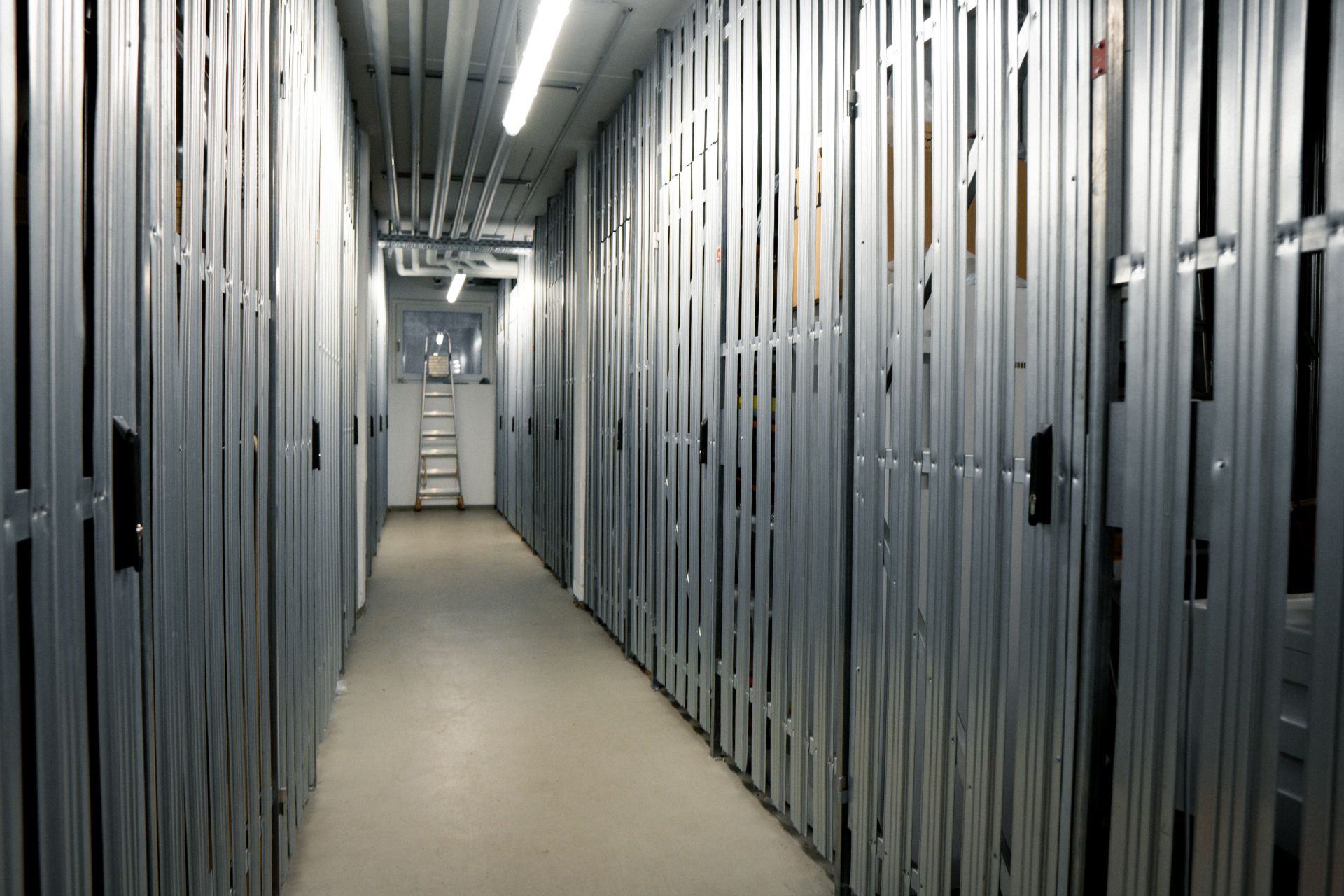 Ein Gang mit metallenen Kellerverschlägen und einer beleuchteten Decke in einem Wohngebäude.
