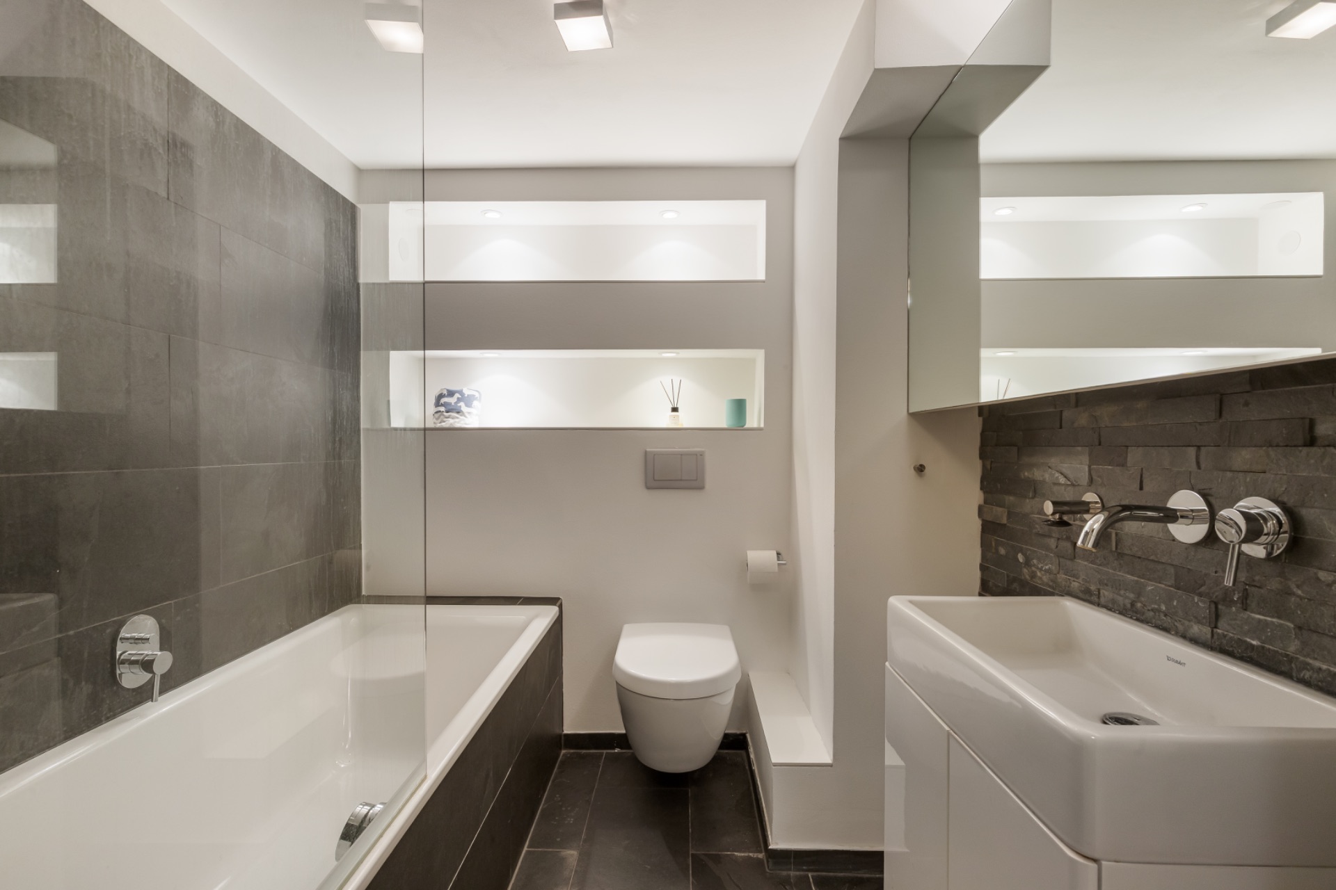 Modernes Badezimmer mit Badewanne, Wandtoilette, Waschbecken und beleuchteten Regalen.