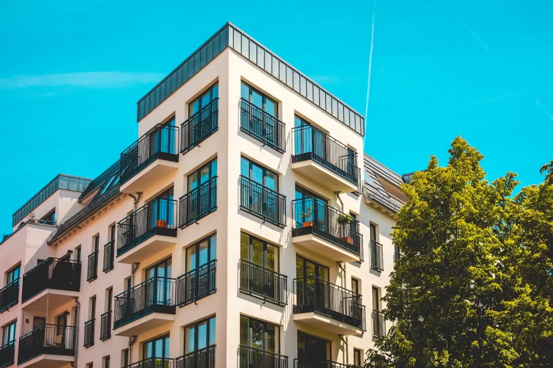 Modernes Wohngebäude mit mehreren Balkonen und großen Fenstern, daneben ein grüner Baum.