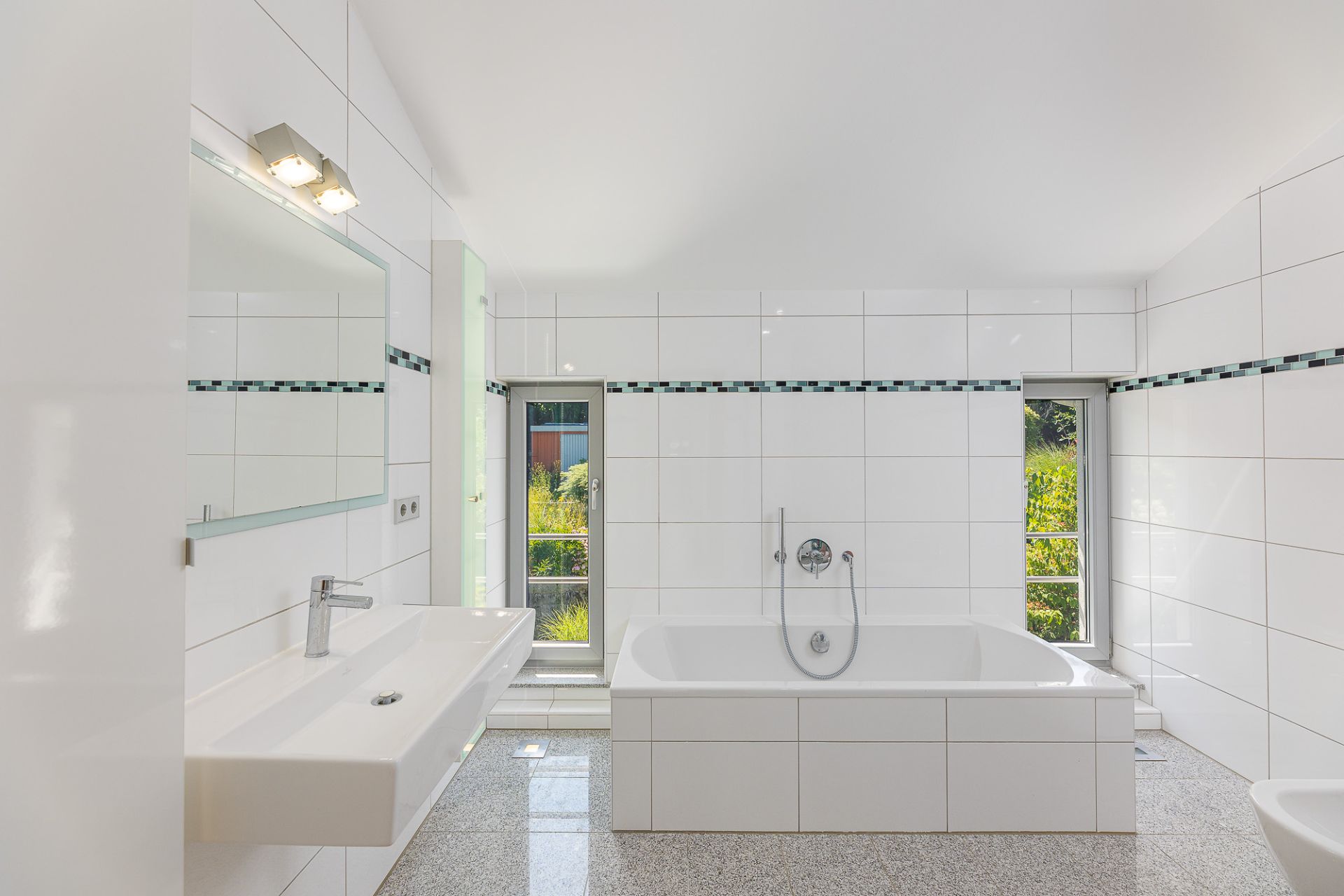 Helles Badezimmer mit weiß gefliesten Wänden, Badewanne, breitem Waschbecken und zwei Fenstern.
