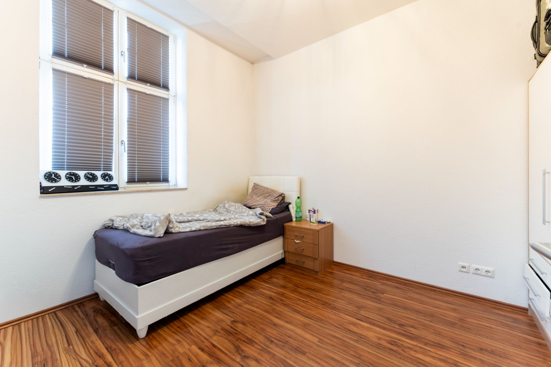 Ein einfach eingerichtetes Schlafzimmer mit Einzelbett, Nachttisch und großem Fenster mit Jalousien.