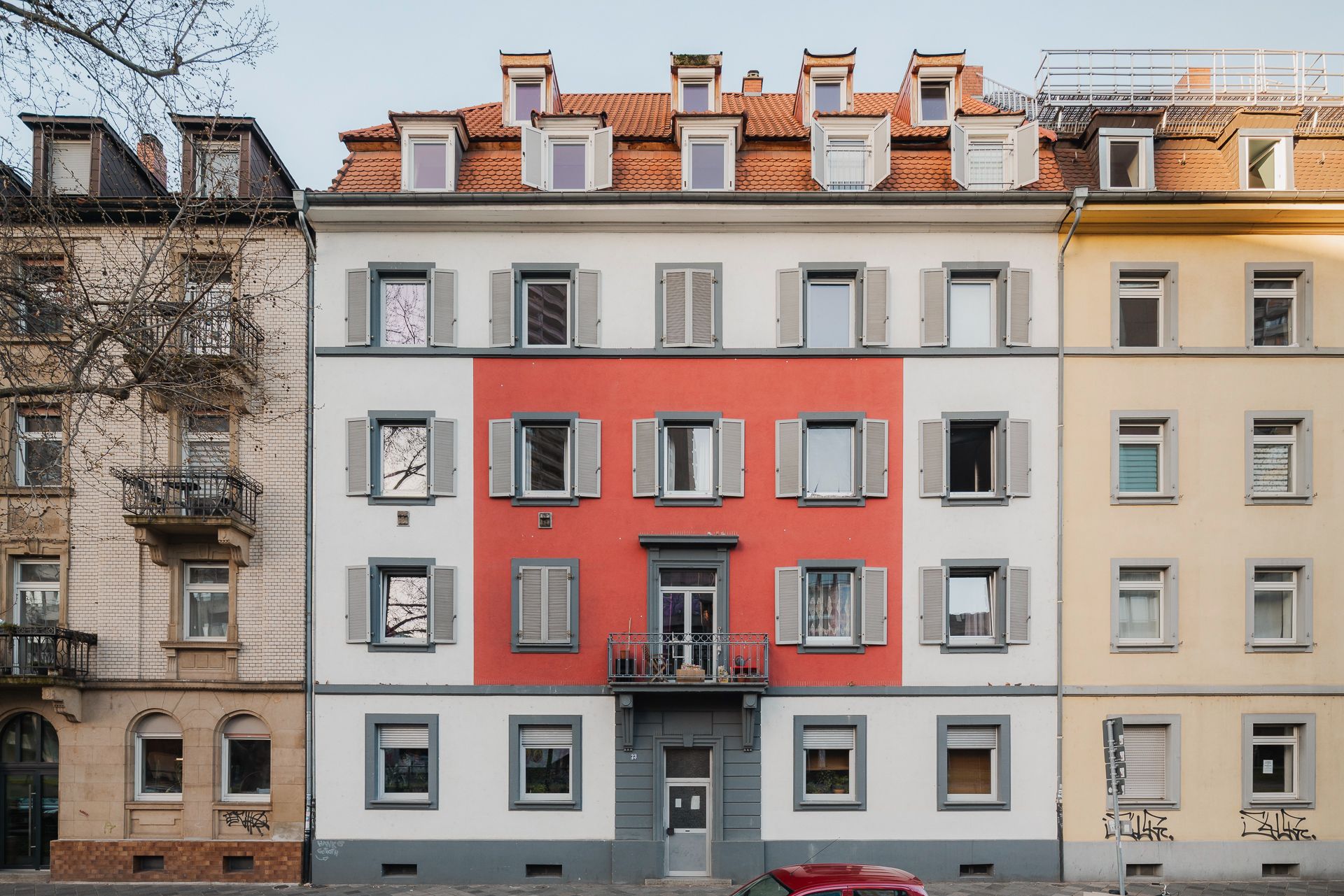 Wohnhaus mit roter Fassade in der Mitte, grauen Fensterläden und kleinem Balkon über dem Eingang.