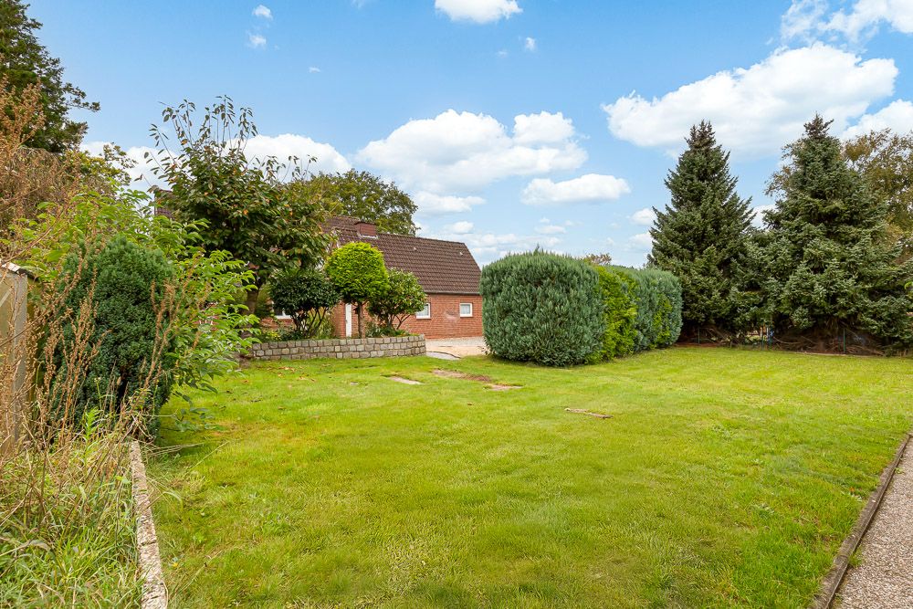 Großer Garten mit Rasen, Bäumen und Büschen, im Hintergrund ein rotes Haus mit braunem Dach.
