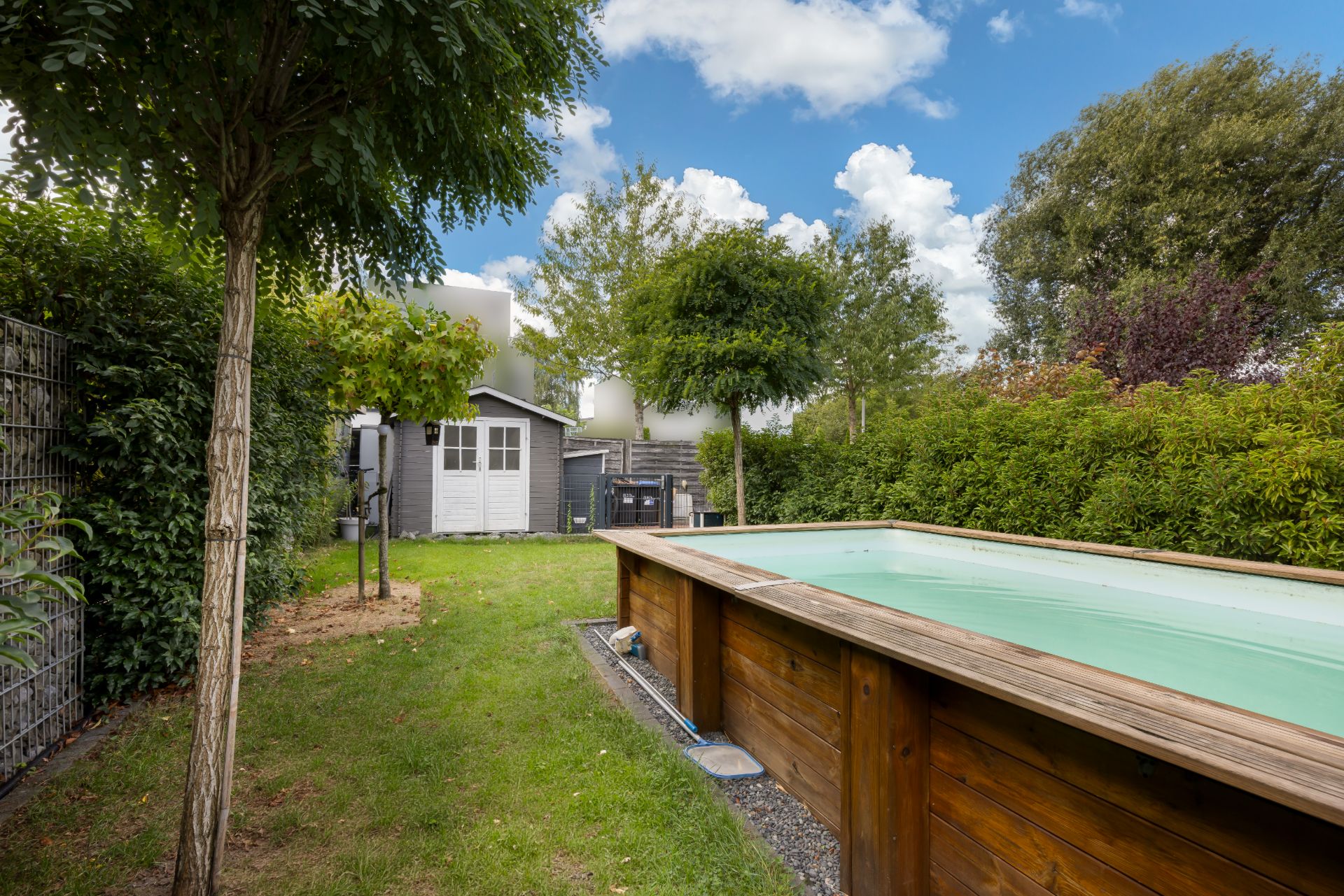Garten mit Holzpool, Bäumen, Rasen und einem kleinen Gartenhaus im Hintergrund.