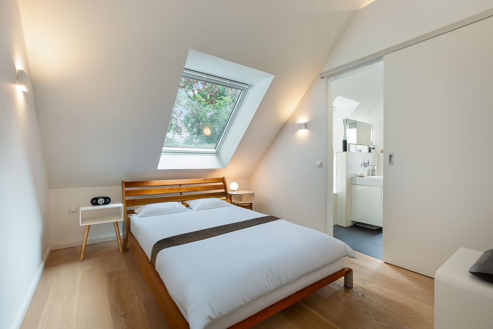 Helles Schlafzimmer mit Dachfenster, Holzbett, Nachttischen und Blick ins Badezimmer.