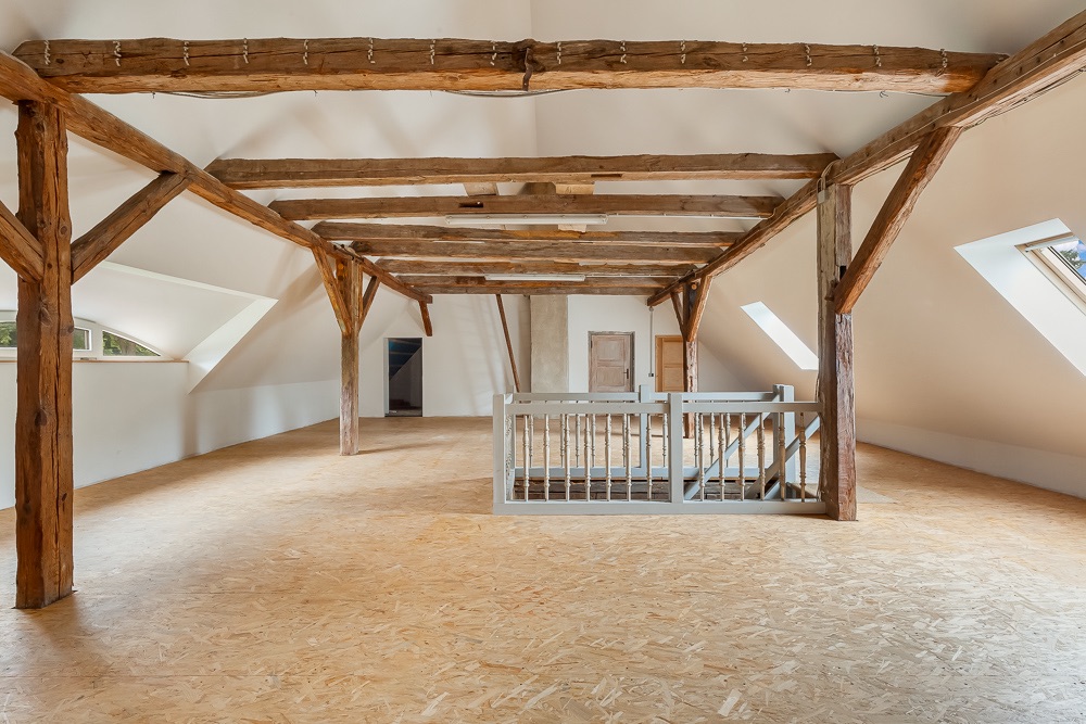 Geräumiges Dachgeschoss mit Holzbalken, offenen Fenstern und Treppengeländer in der Mitte.