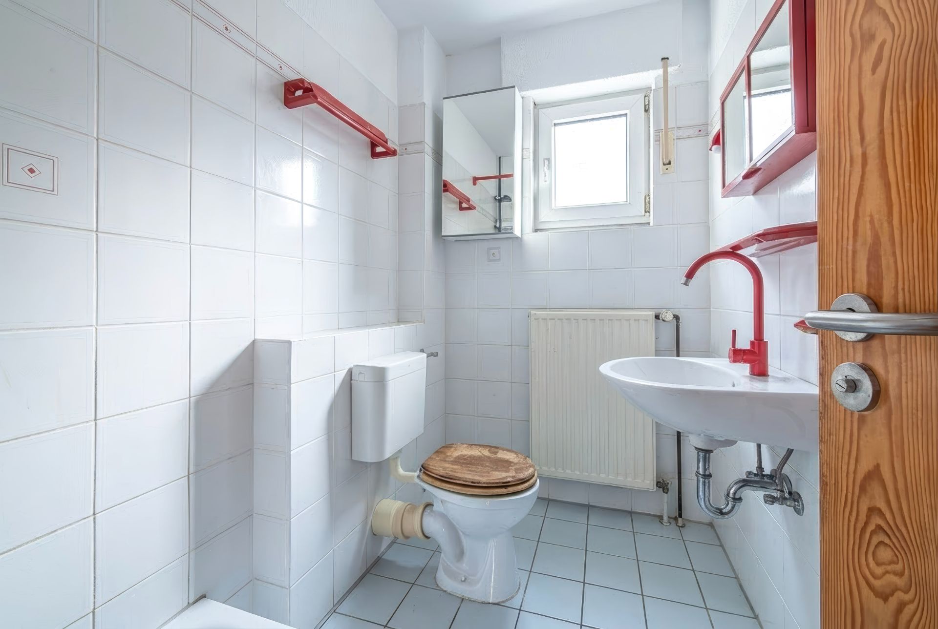 Helles Badezimmer mit WC, Waschbecken, Spiegel, Fenster und roten Ablagen.