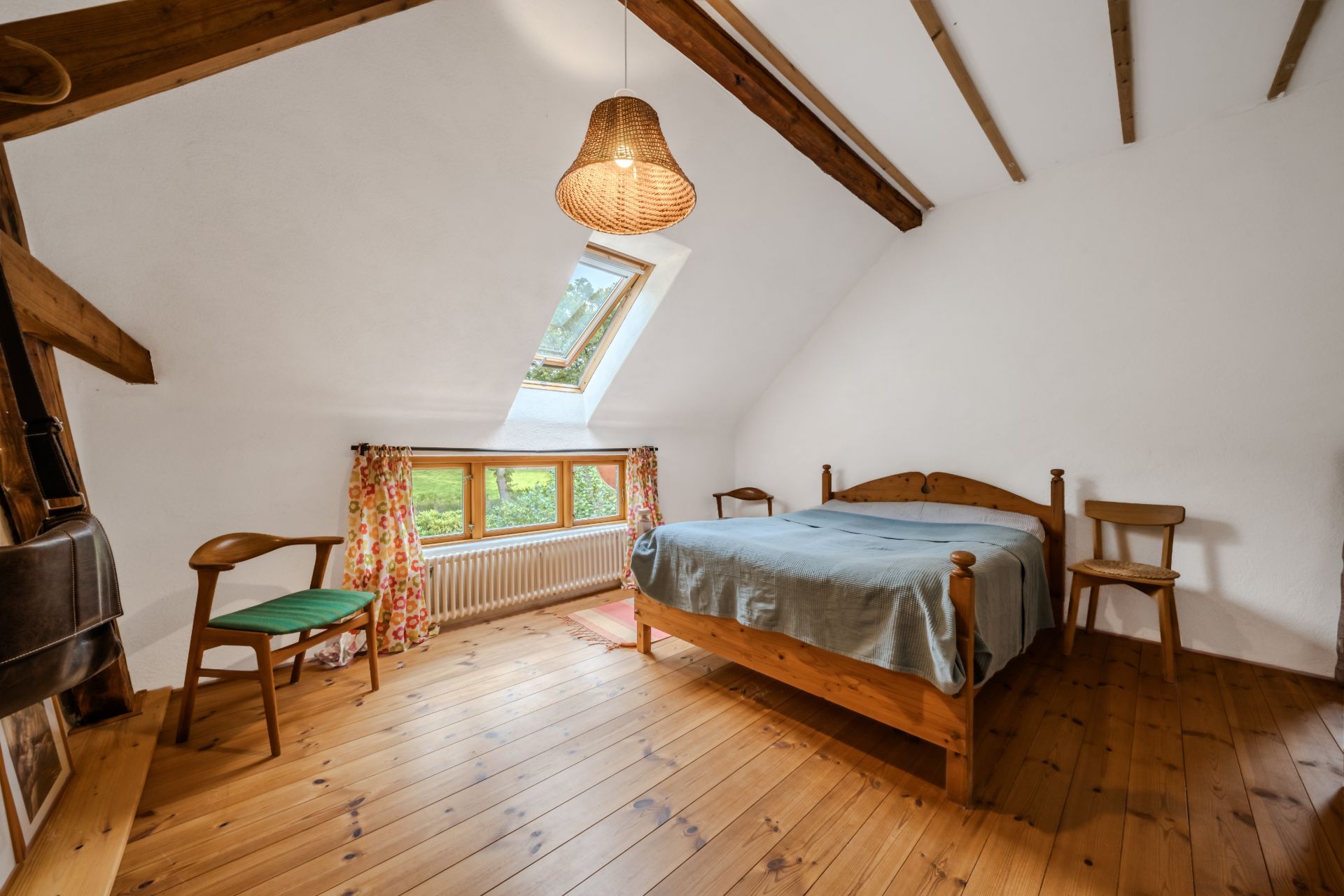 Schlafzimmer mit Holzboden, Holzbett, Stühlen, Fenster mit Vorhängen und Dachfenster.