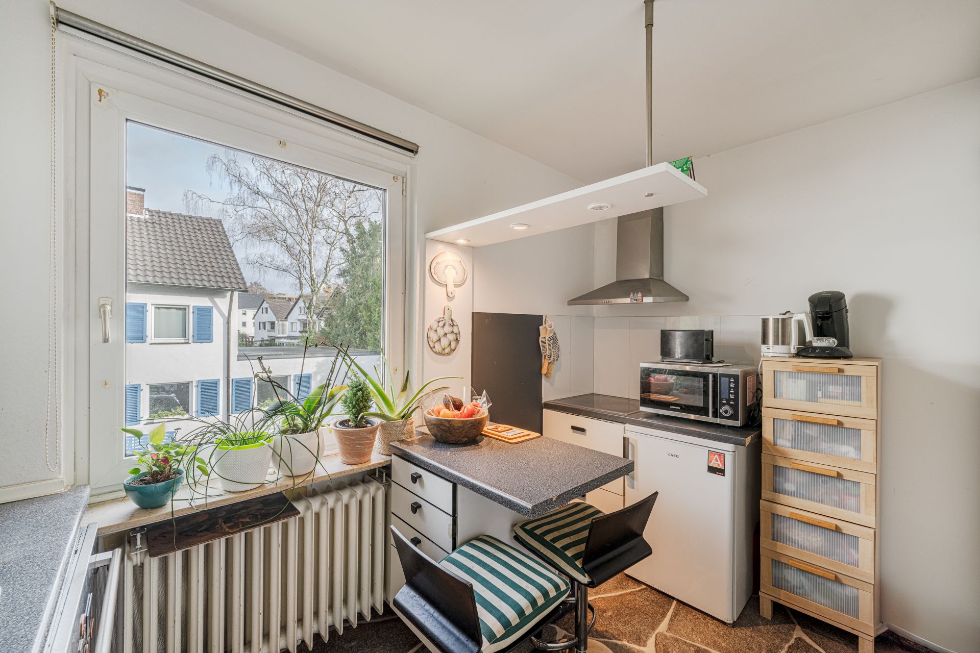 Kleine Küche mit Esstisch, zwei Stühlen, Pflanzen am Fenster und Haushaltsgeräten.