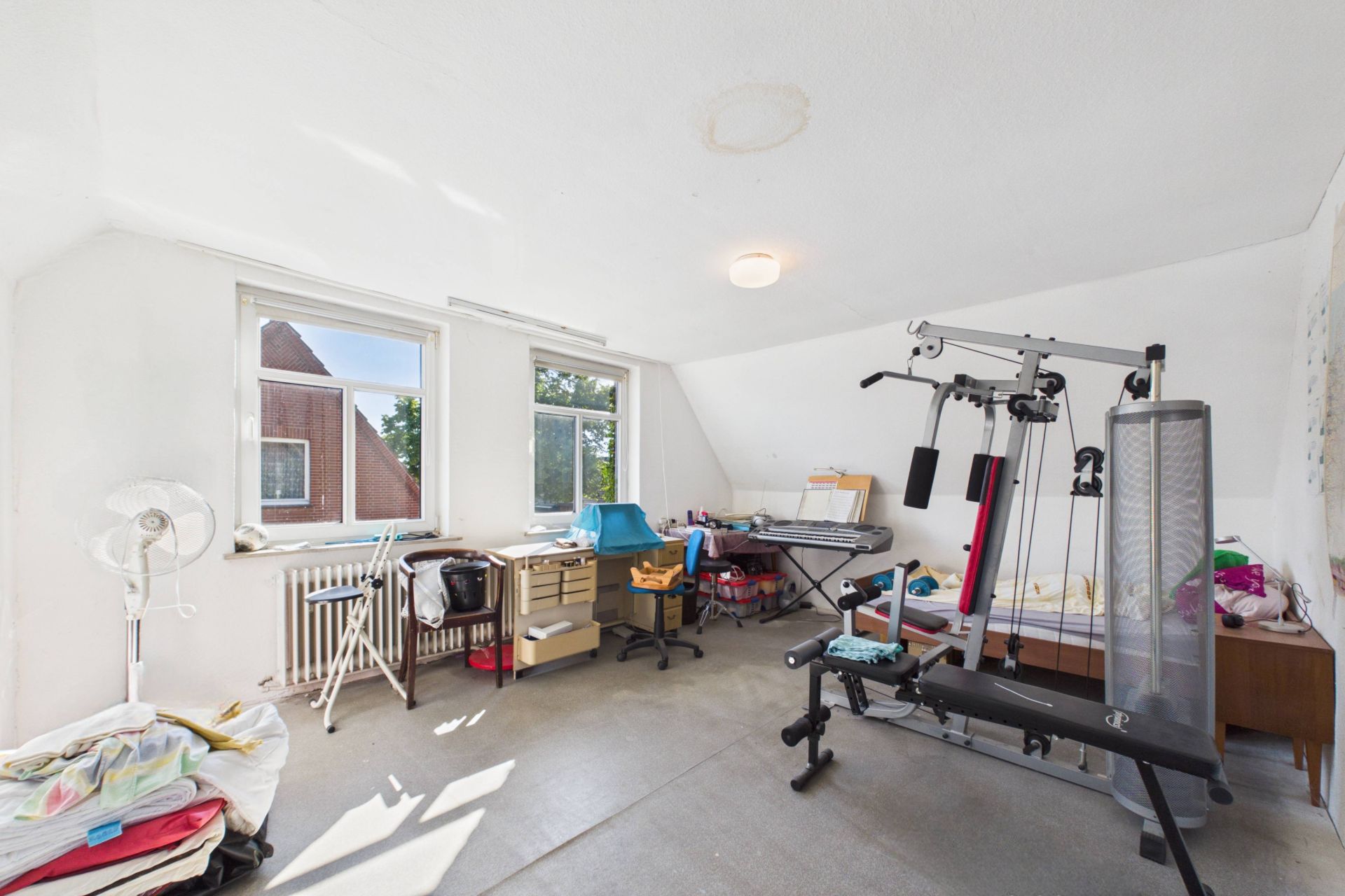 Helles Zimmer mit zwei Schreibtischen, Fitnessgerät und Wäsche auf einem Stuhl.