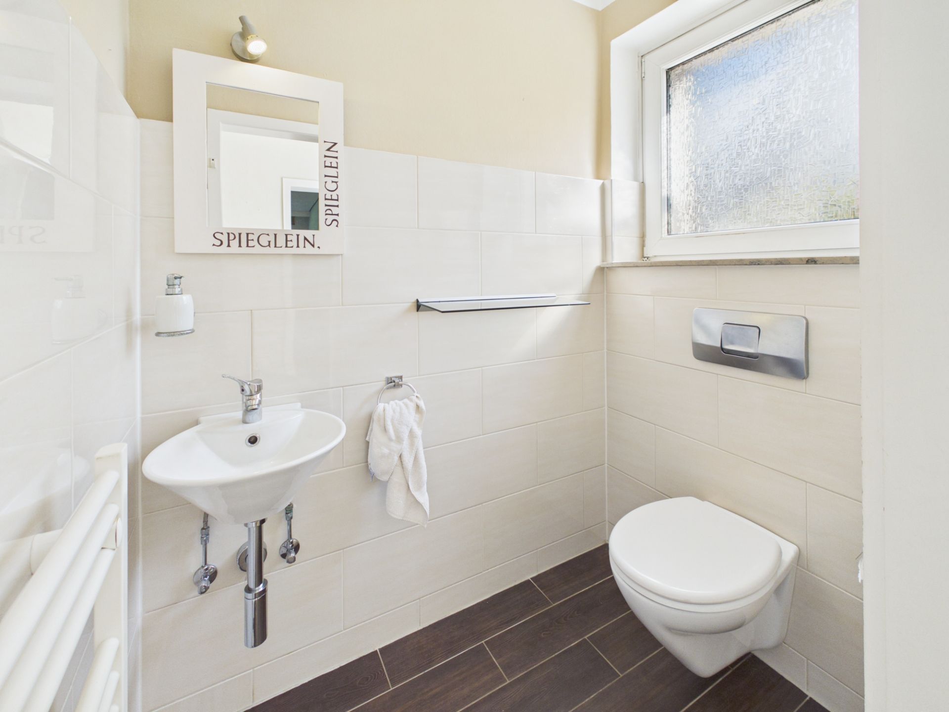 Helles Badezimmer mit kleinem Waschbecken, Spiegel, WC und Fenster.