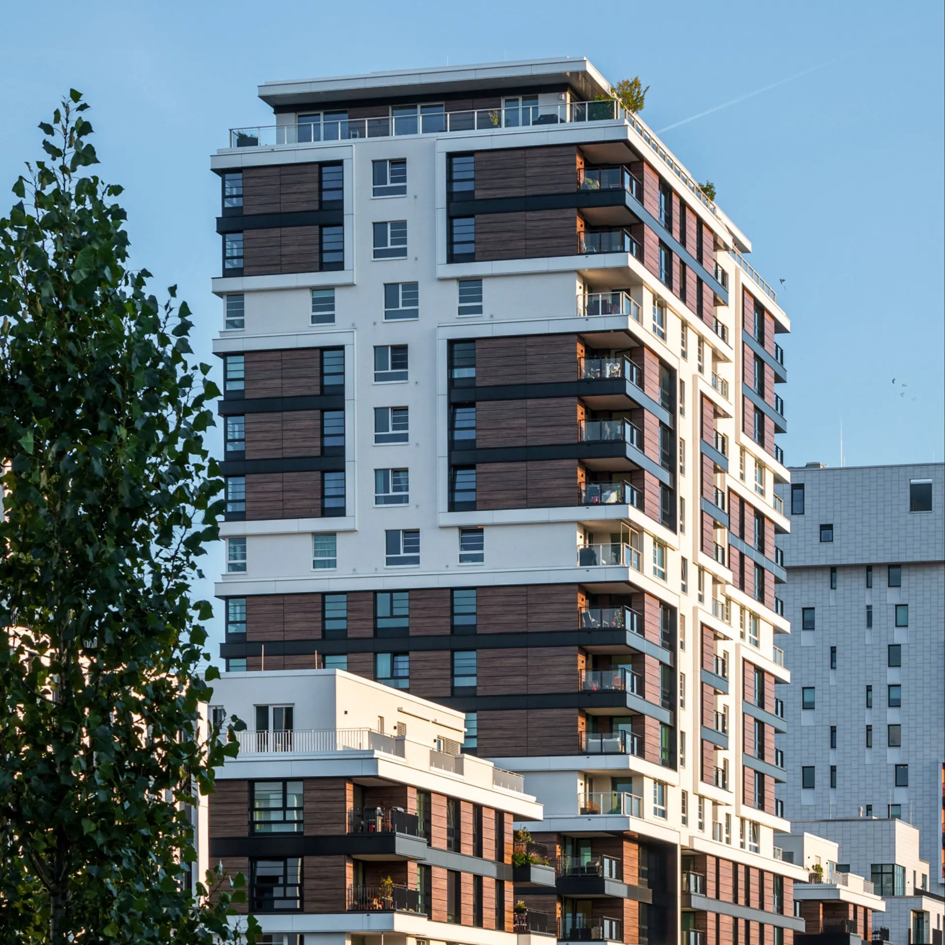Modernes Wohnhaus mit mehreren Etagen und Balkonen, Bäume im Vordergrund.