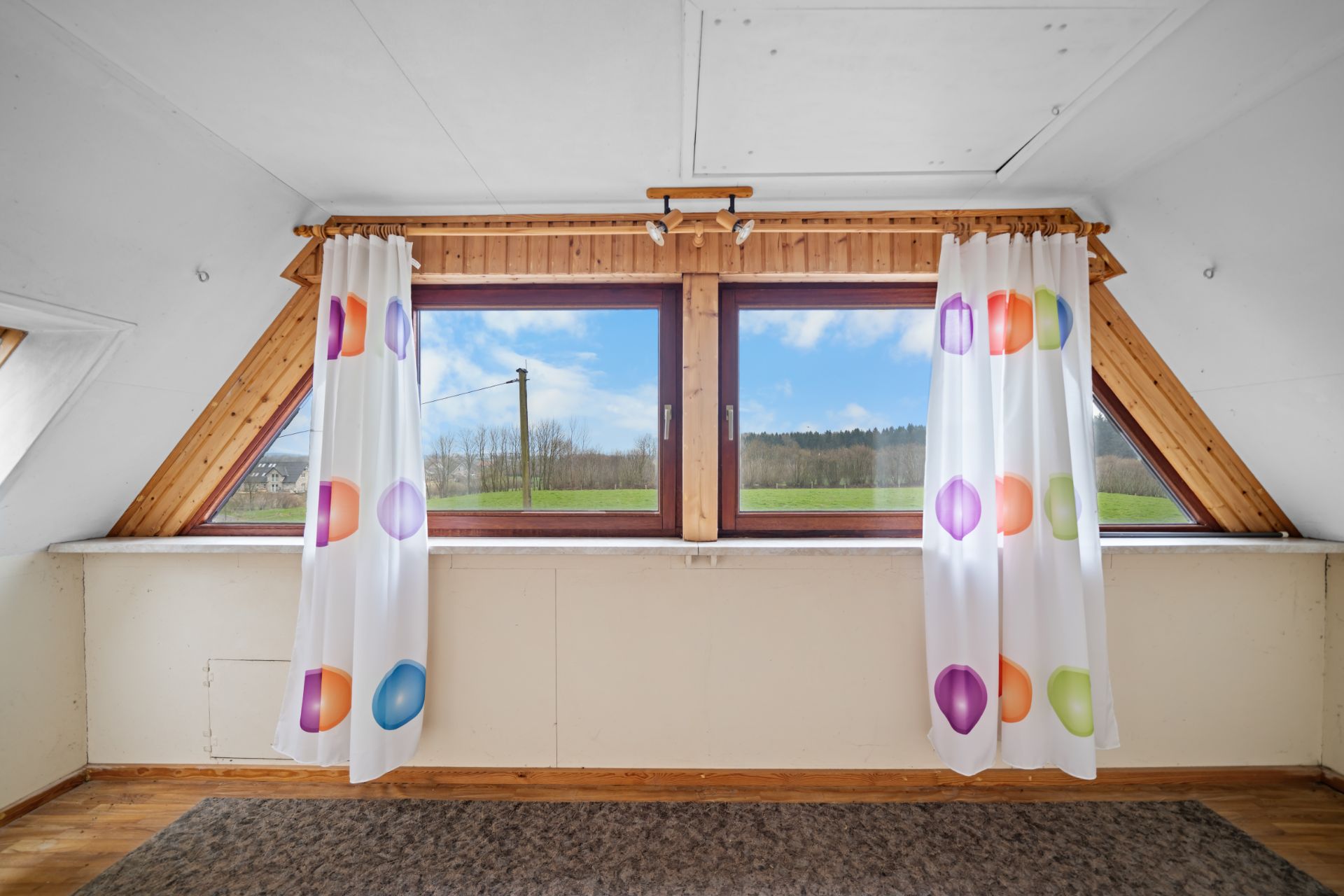 Fensterfront mit bunten Vorhängen und Blick auf grüne Wiese und blauen Himmel.