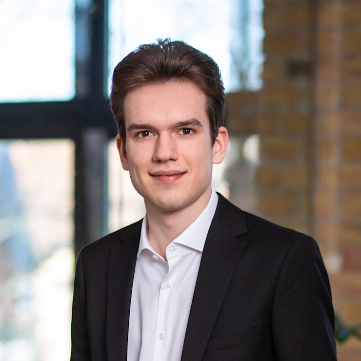 Junger Mann im Anzug steht vor einem Fenster mit Backsteinwand im Hintergrund.